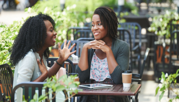 Pretty African-American woman having coffee and telling story to her female friend at a restaurant on a bright sunny day