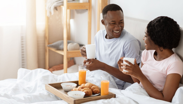 Joyful black young couple sitting in bed in morning drinking coffee and talking with breakfast in bed